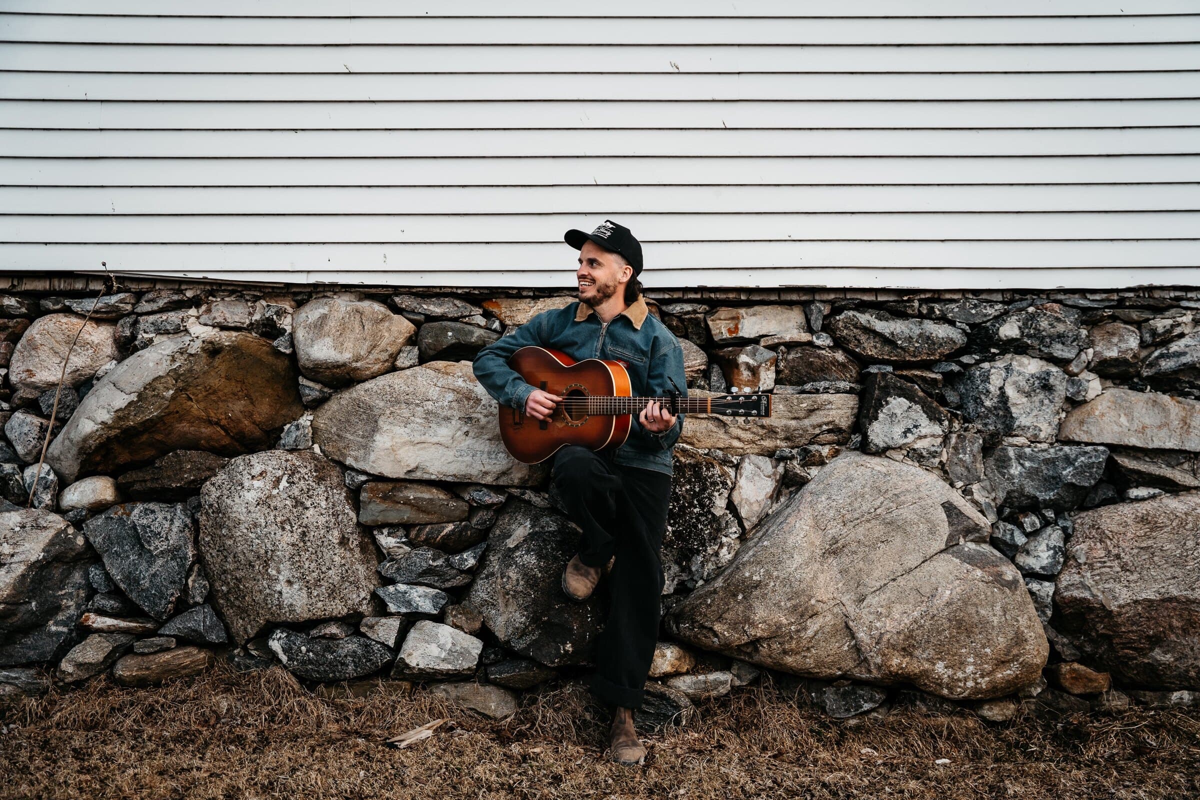Joseph Poirier, souriant, joue de la guitare assis sur un muret de pierres au pied d'une maison blanche