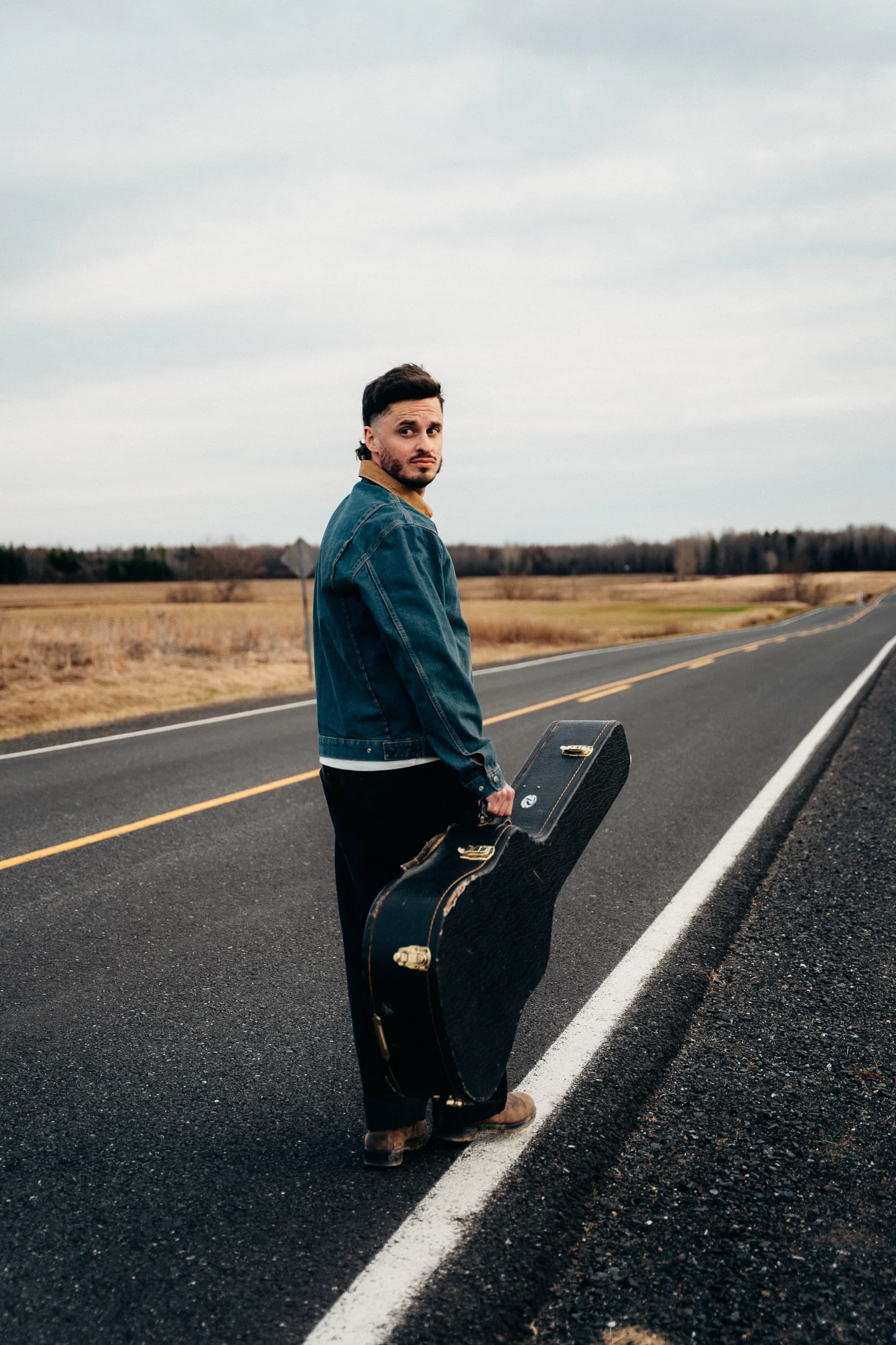 Joseph Poirier sur une route de campagne, un étui de guitare à la main, qui se retourne vers nous